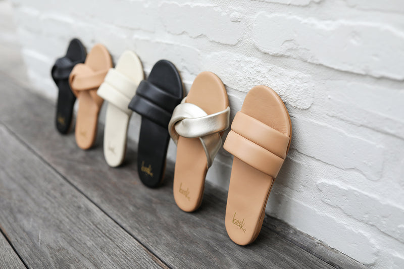 Group shot of Twistybird leather sandals in black, beach, platinum/beach and Sugarbird slide sandals in eggshell, black, and beach.