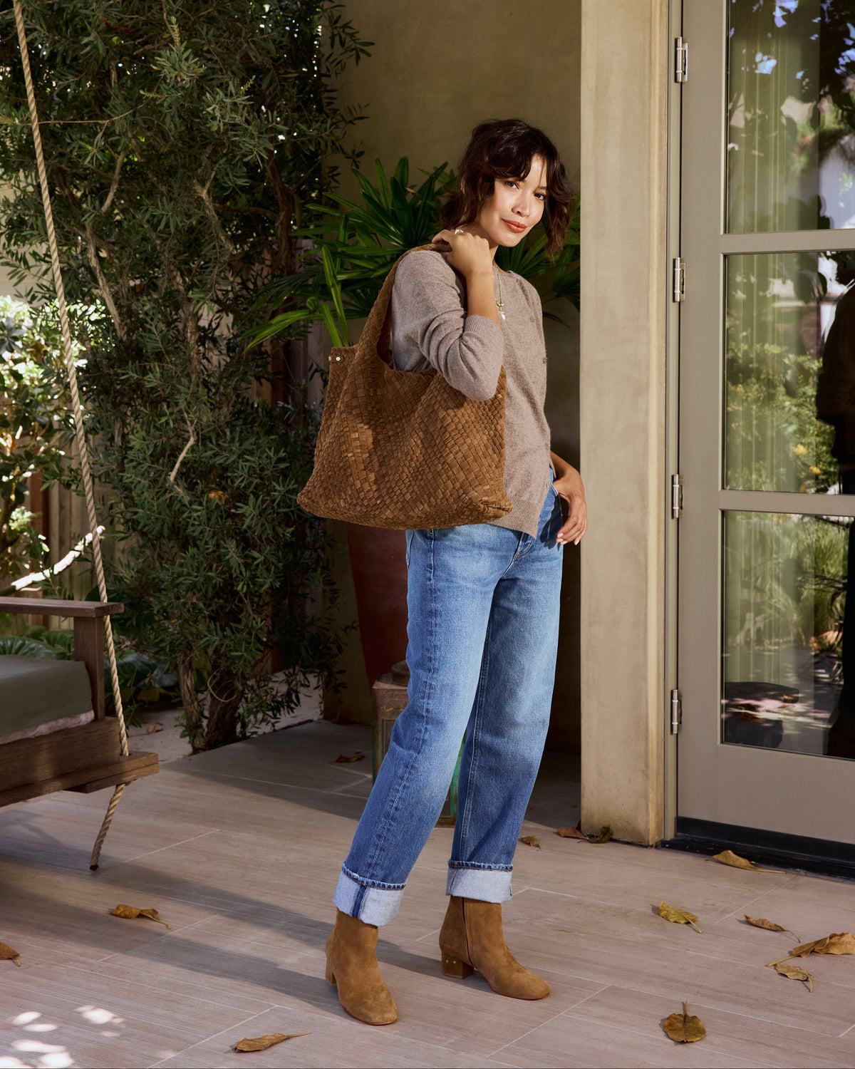A woman wearing blue jeans , sweater, and Raven suede boots in toffee carries the Willet suede woven tote bag in chestnut in an outdoor setting.