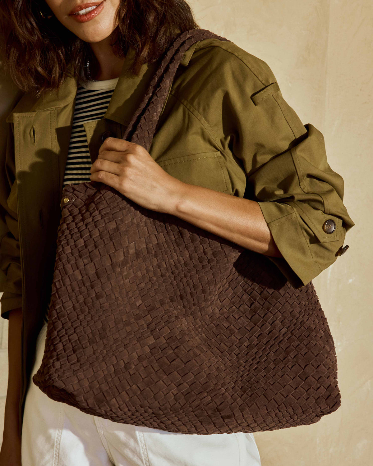 A woman wearing an olive green jacket, a black and white striped shirt, white pants carries the Willet suede woven tote bag in chocolate