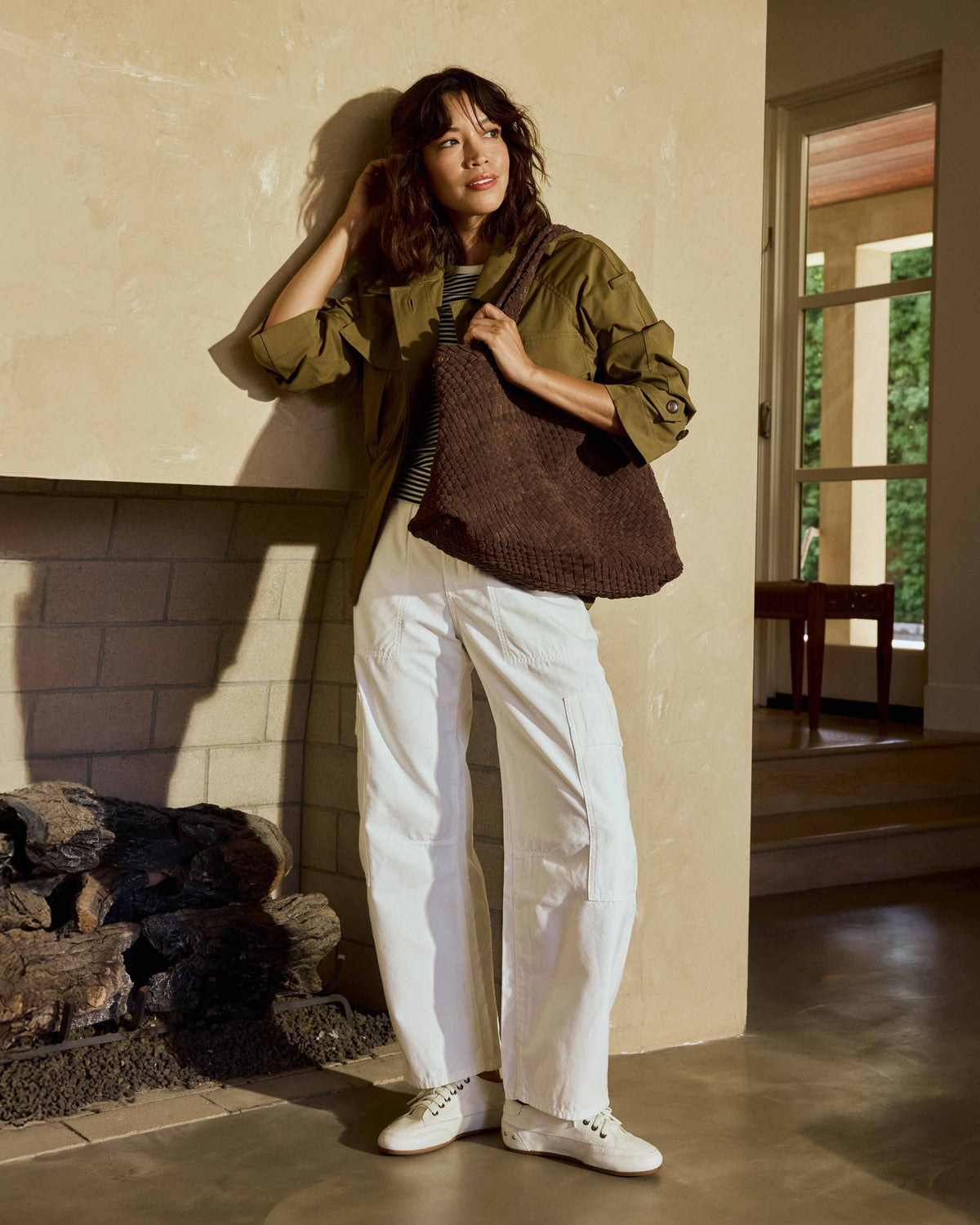 A woman wearing an olive green jacket, a black and white striped shirt, white pants, and Sandpiper leather sneakers in white carries the Willet suede woven tote bag in chocolate