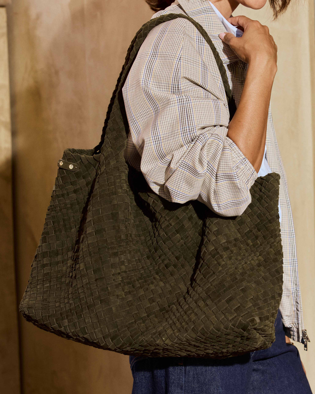 A woman wearing a beige plaid shirt and blue jeans carries the Willet suede woven tote bag in olive
