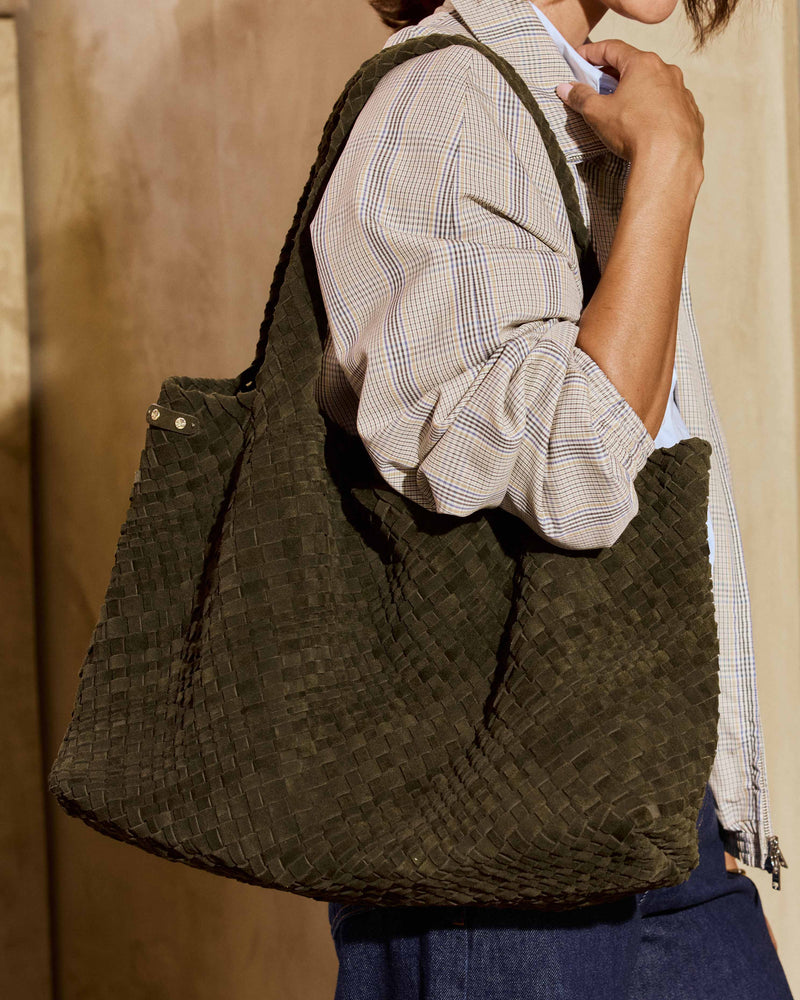 A woman wearing a beige plaid shirt and blue jeans carries the Willet suede woven tote bag in olive