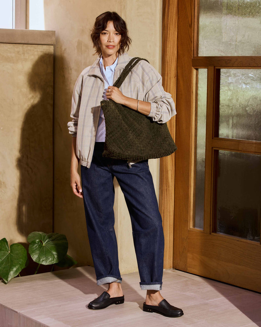 A woman wearing a beige plaid shirt, blue jeans, Owlet leather loafer mules in black carries the Willet suede woven tote bag in olive