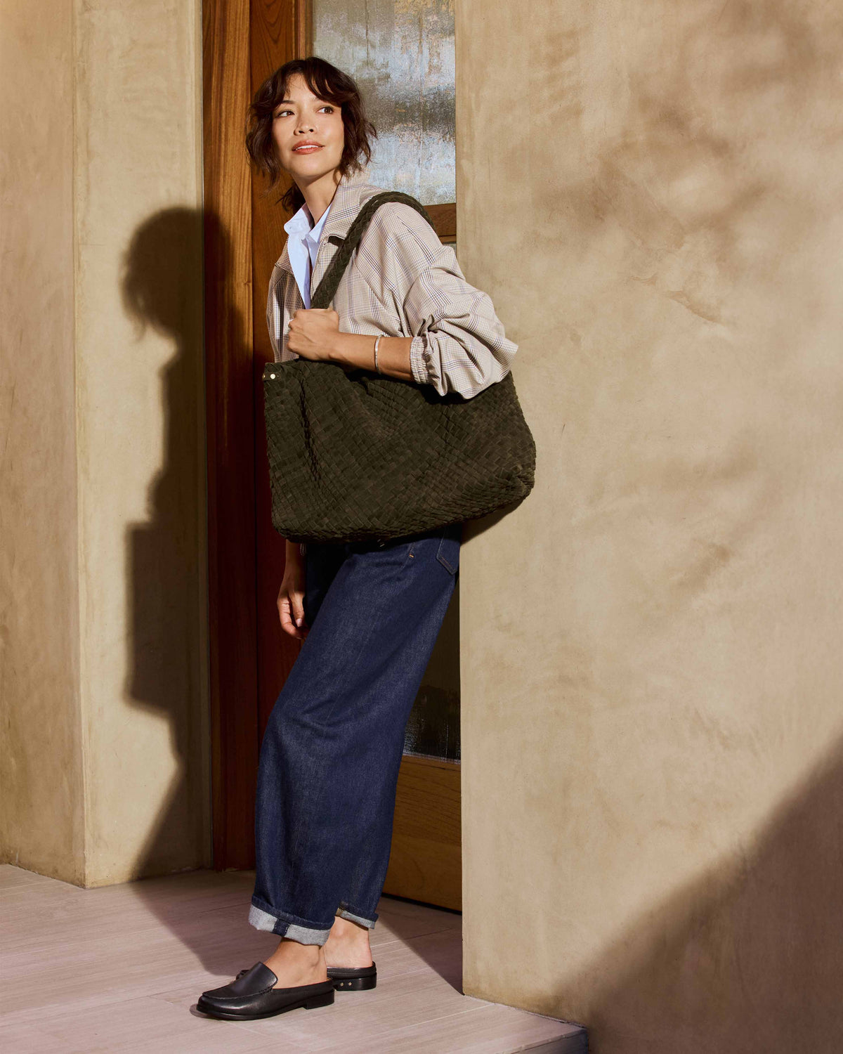 A woman wearing a beige plaid shirt, blue jeans, Owlet leather loafer mules in black carries the Willet suede woven tote bag in olive