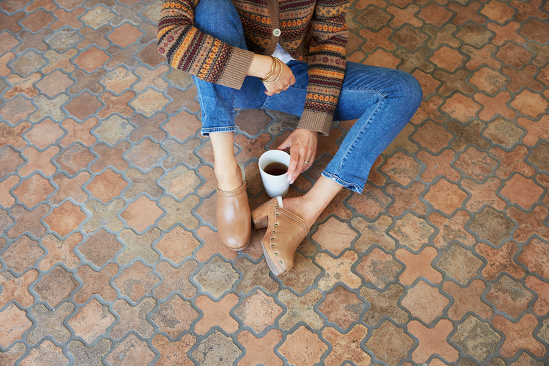 Woman wearing Woodpecker Más Shearling clogs in saddle with jeans and sweater, holding coffee cup.