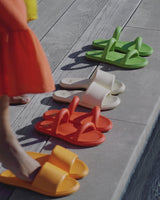 Woman wearing Puffbird slide sandals in papaya, eggshell, and beach. Woman wearing Pintail slide sandals in flamingo and leaf.