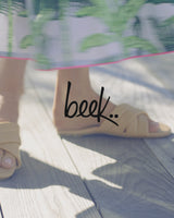 Woman wearing Surfbird leather slide sandals in beach. Group shot of Surfbird leather slide sandals in leaf, poppy, eggshell, beach and Sugarbird leather slide sandals in lilac, papaya, platinum/beach.