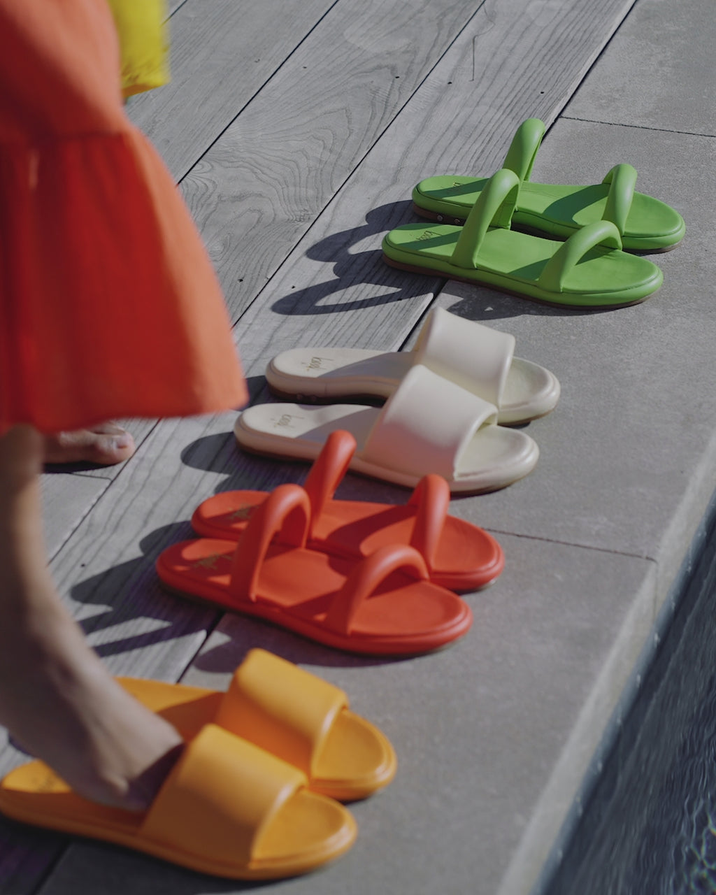 Woman wearing Puffbird slide sandals in papaya, eggshell, and beach. Woman wearing Pintail slide sandals in flamingo and leaf.